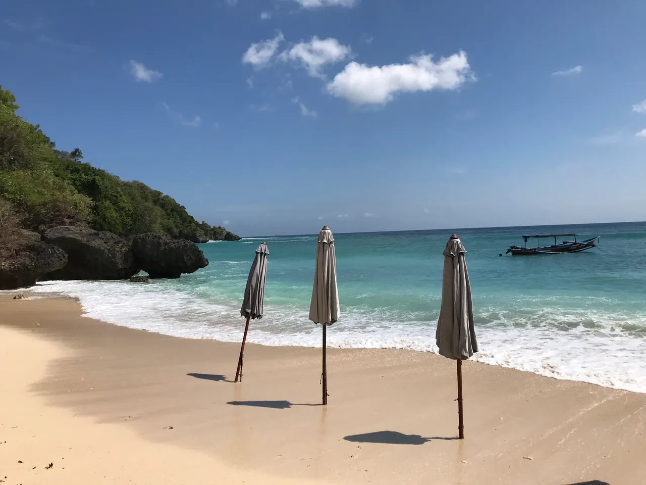 Soft sand, closed parasols and a traditional outrigger boat on turquoise water at Padang Padang Beach, Bali.
