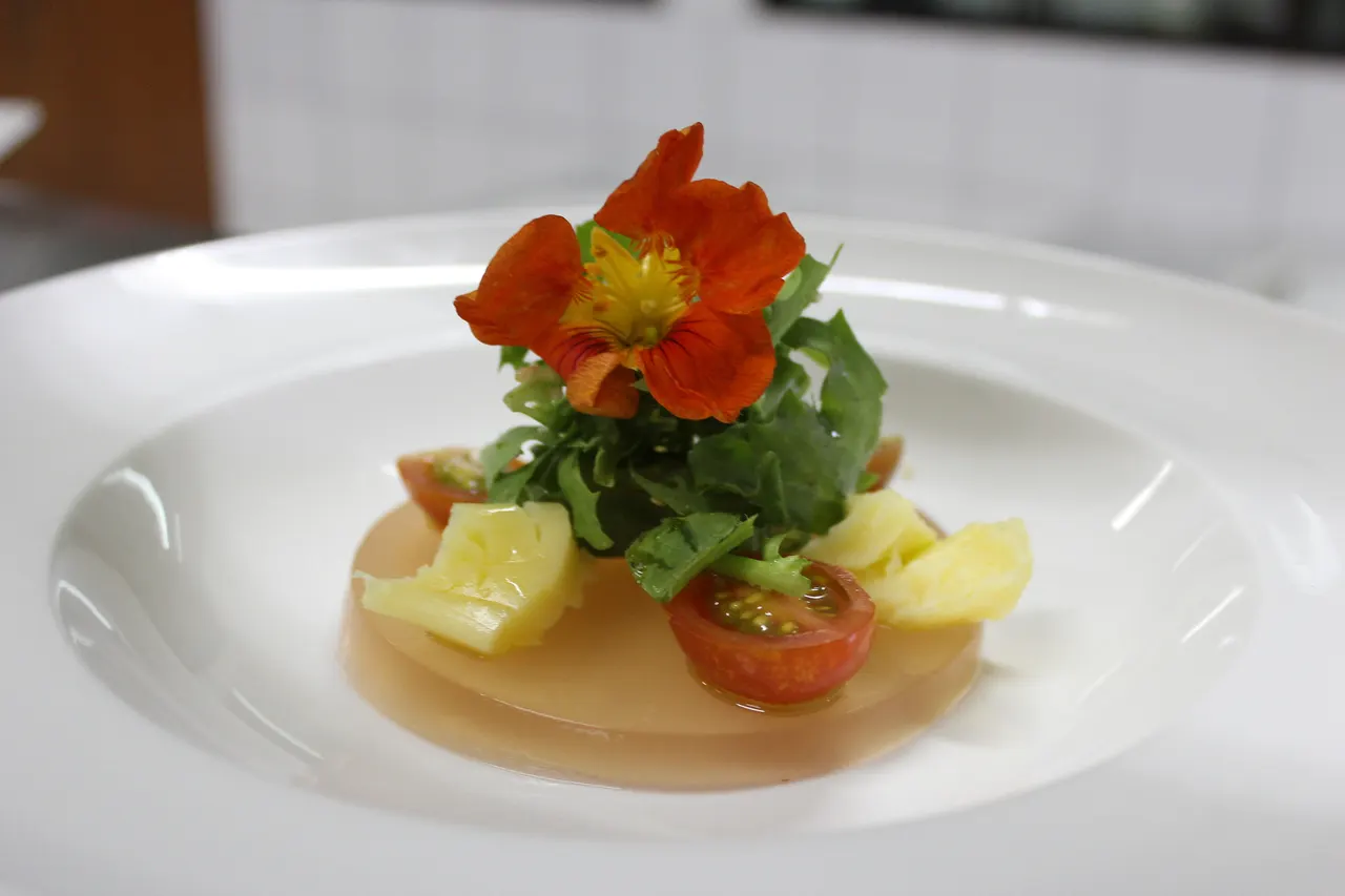 Fine dining appetizer on a white plate: nasturtium flower, rocket leaves, cherry tomato and pineapple.