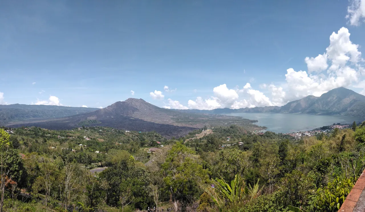 Mount Batur volcano and its black lava field above Lake Batur, seen from the Kintamani ridge in Bali under a blue sky.